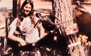 Black and white photograph of a caucasian woman with long blonde hair, in a white shirt with sunglasses on her head, leaning on a black motorcycle by a tree. Photographed during the day