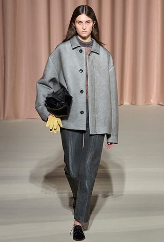 a photo showing the fall jacket trend, chore jackets on the Philosophy di Lorenzo Serafini runway with a model wearing a gray coat over a gray knit with gray trousers, black oxfords, a black bag, and yellow leather gloves