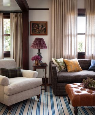 Traditional interior with dark wood features, windows dressed with sheer drapes, white painted walls, blue and white striped rug, brown sofa, cream armchair, leather ottoman