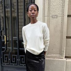 Influencer @sylviemus_ stands outside in Paris wearing a cream fine-knit jumper with a black sequinned pencil skirt and pointed-toe heels.