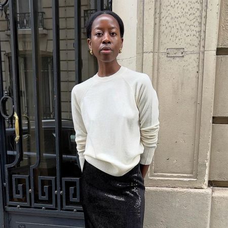 Influencer @sylviemus_ stands outside in Paris wearing a cream fine-knit jumper with a black sequinned pencil skirt and pointed-toe heels.