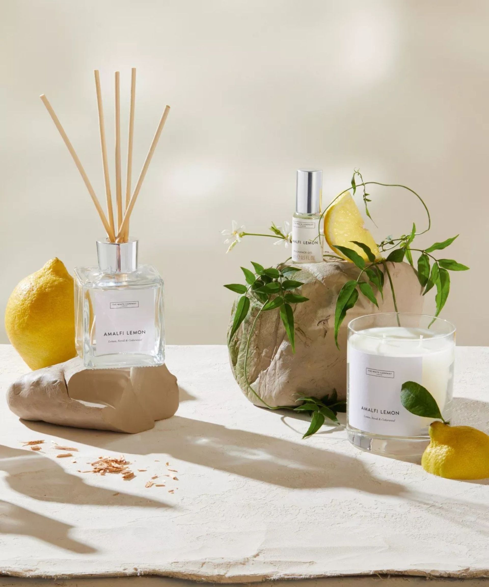 Reed diffuser, spray bottle and candle on a white surface surrounded by lemons and green leaves.