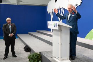 Boris Johnson wearing a suit and tie standing below a podium where King Charles is raising his arms and speaking enthusiastically