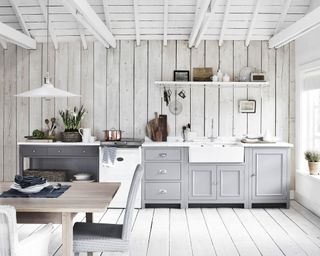 White kitchen spaces with white wooden ceiling, off-white wooden paneled walls, white wooden flooring, gray kitchen units, white hanging pendant, wooden dining table with upholstered dining chairs
