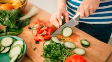 Shot of person's hands chopping cucumbers with a chef's knife on a wooden chopping board. Other veg is in shot