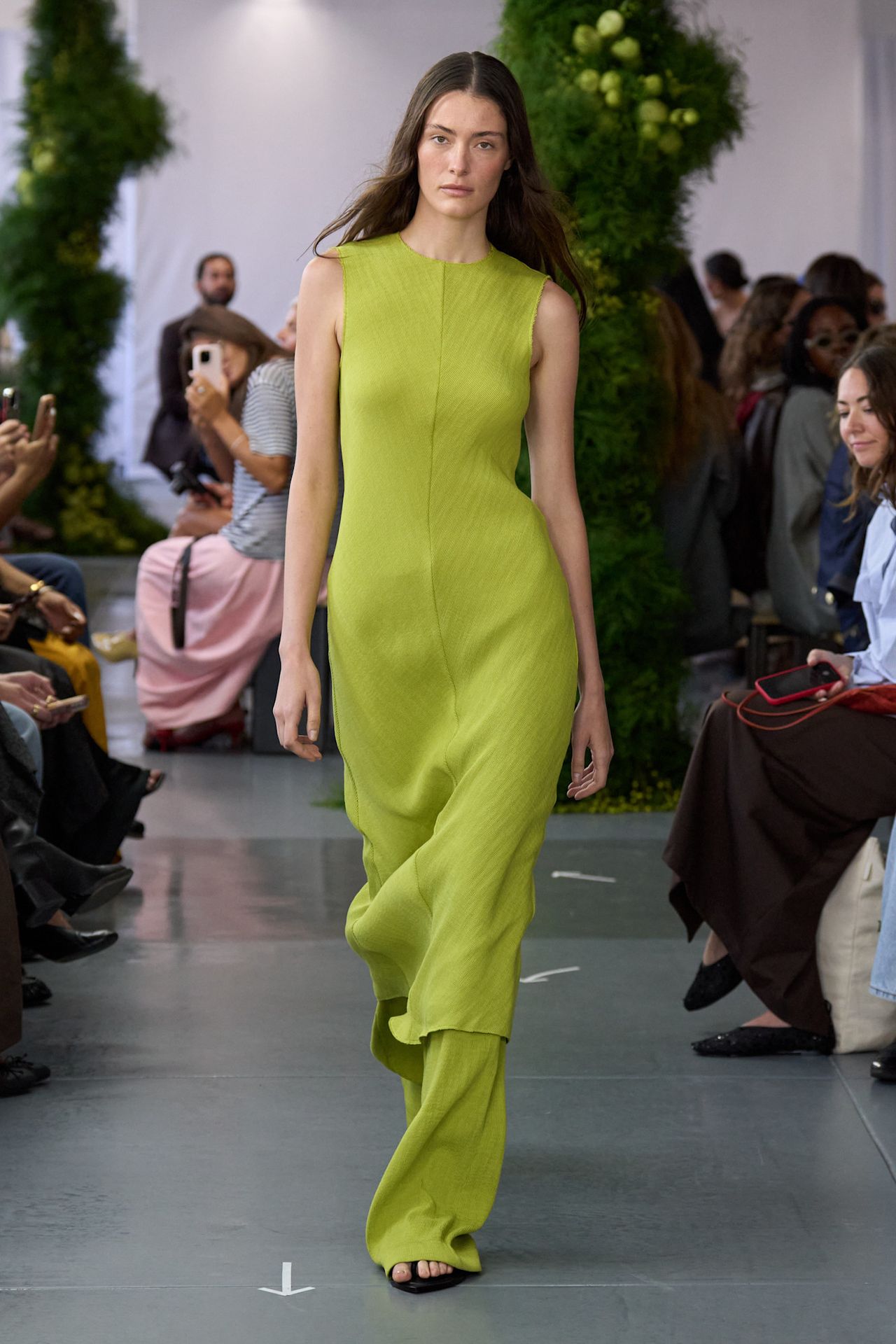 A Tibi model wearing a bright lime green pleated midi dress styled with matching wide-leg pants and black flat sandals.