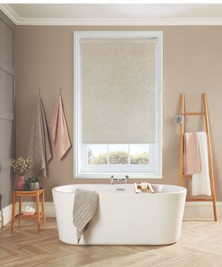 Freestanding white bath on wooden bathroom floor with neutral roller blind on central window in foregrounds. Peach, gray and taupe towels and wooden rails around the bathroom can be seen