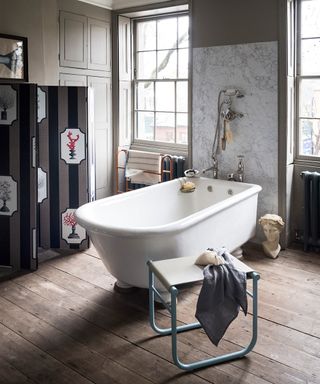 A white bath with one end attached to a wall in between two large windows in a bathroom with wooden floor boards