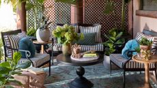 outdoor living room with a large rug, three matching armchairs with striped cushions and a black coffee table all styled with books, plants, florals, and pillows
