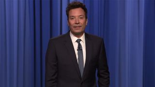 Jimmy Fallon stands in front of a blue curtain in a suit while giving his monologue on The Tonight Show.