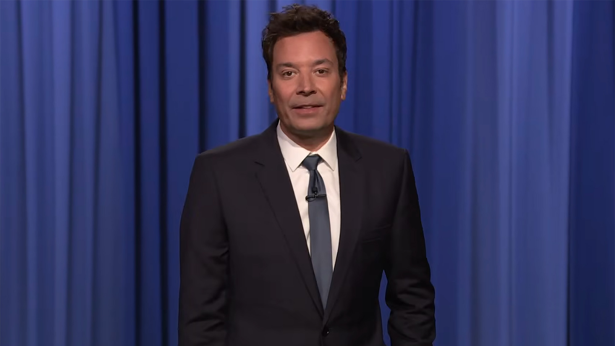 Jimmy Fallon stands in front of a blue curtain in a suit while giving his monologue on The Tonight Show.