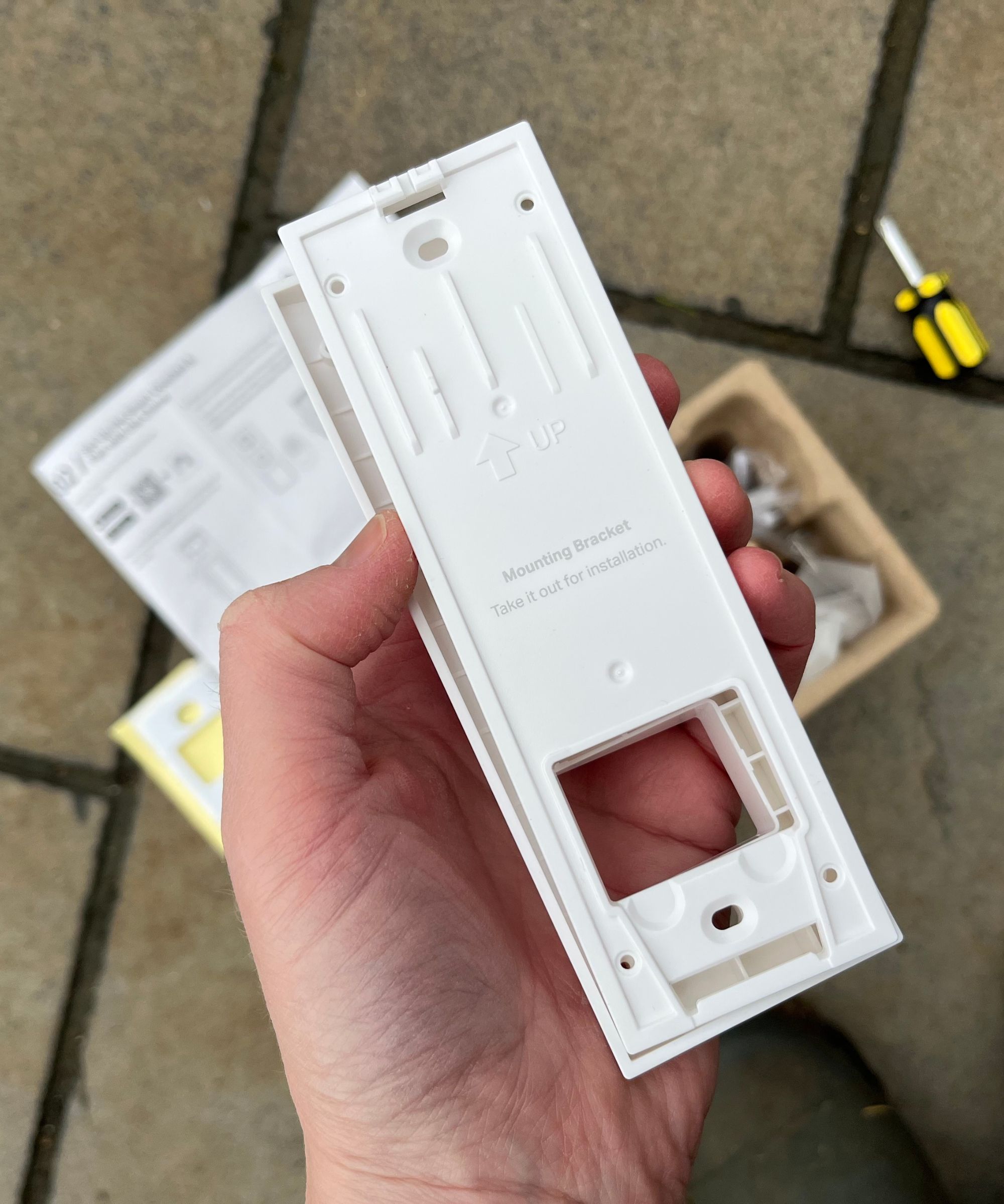 Hand holding the back plate of the Tapo D210 Video Doorbell above other parts and tiled concrete floor.
