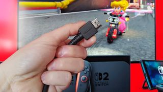 Hand holding Nintendo branded HDMI cable with Switch 2 promotional material on screen in backdrop with Peach in Mario Kart World on motorbike.