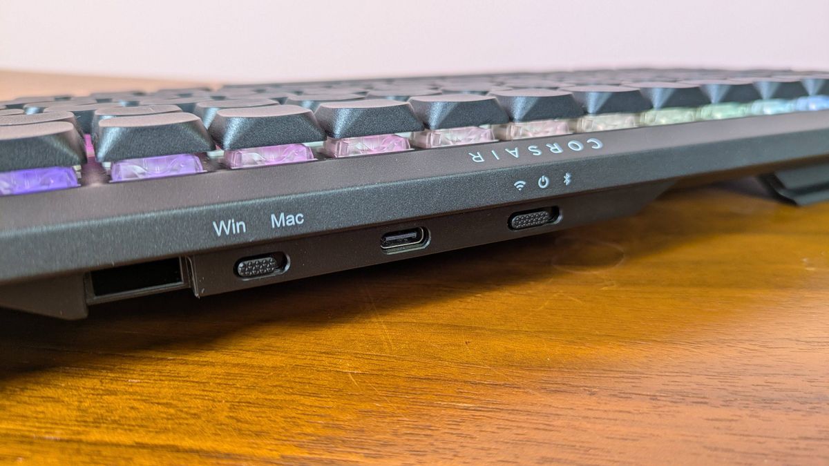 Close-up of rear switches and USB-C port on Corsair Vanguard Air 99 Wireless, on a brown desk with pink wall in background