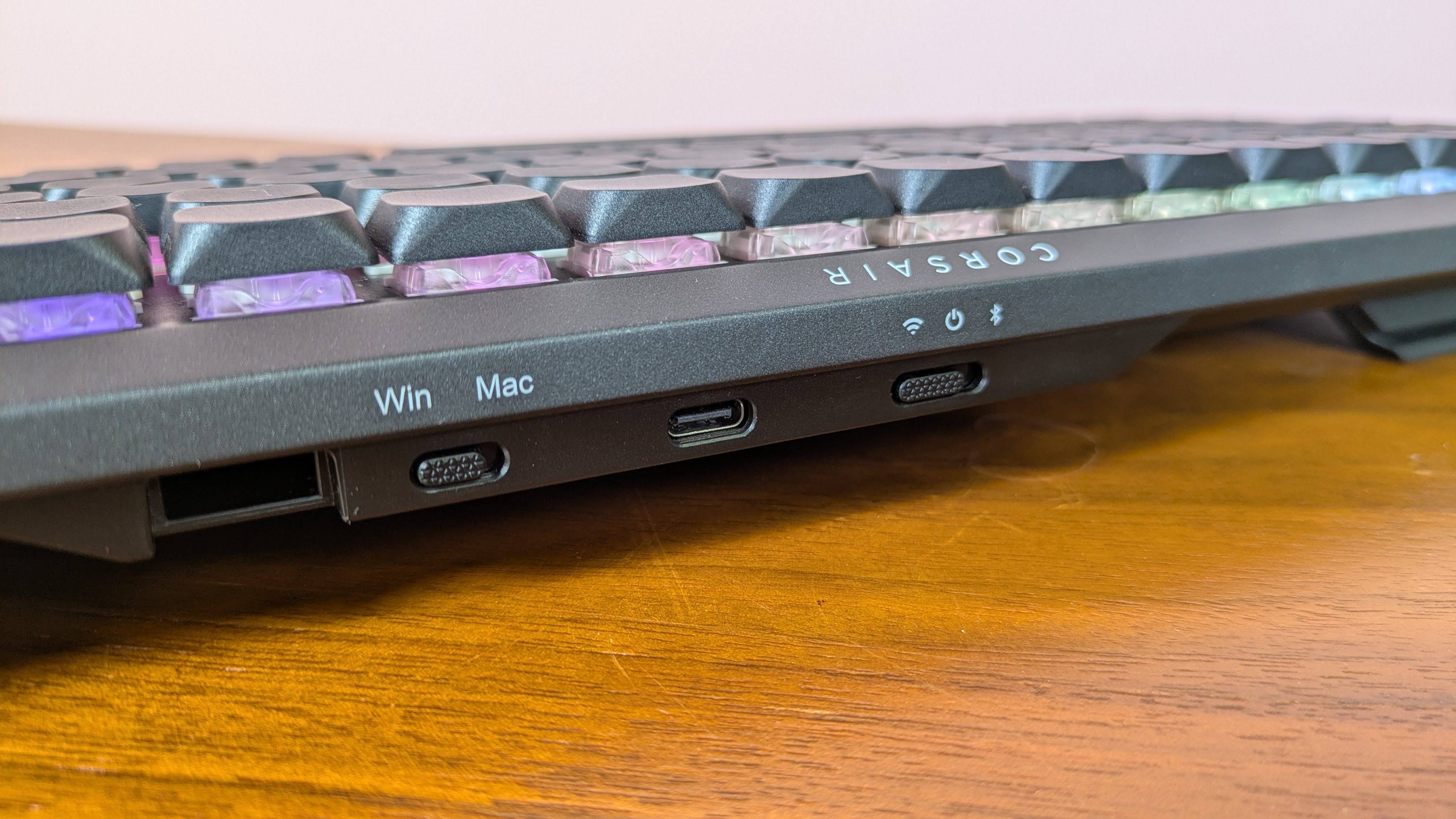 Close-up of rear switches and USB-C port on Corsair Vanguard Air 99 Wireless, on a brown desk with pink wall in background