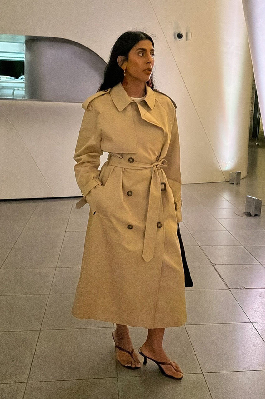 A woman wearing a tan trench coat and black kitten heel thong sandals.