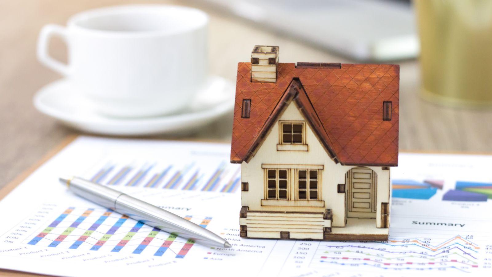A small wooden house model sits on financial charts beside a silver pen and white coffee cup, suggesting property investment or real estate market analysis.