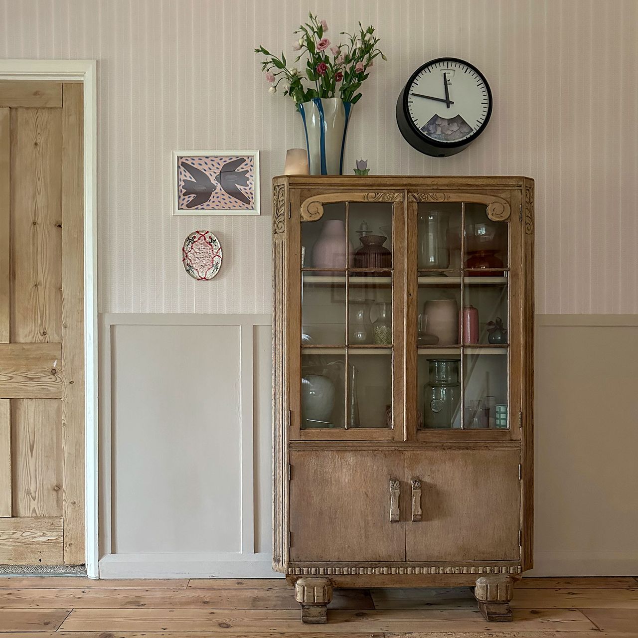 Neutral hallway with wall panelling and wooden glass fronted cabinet storing vases