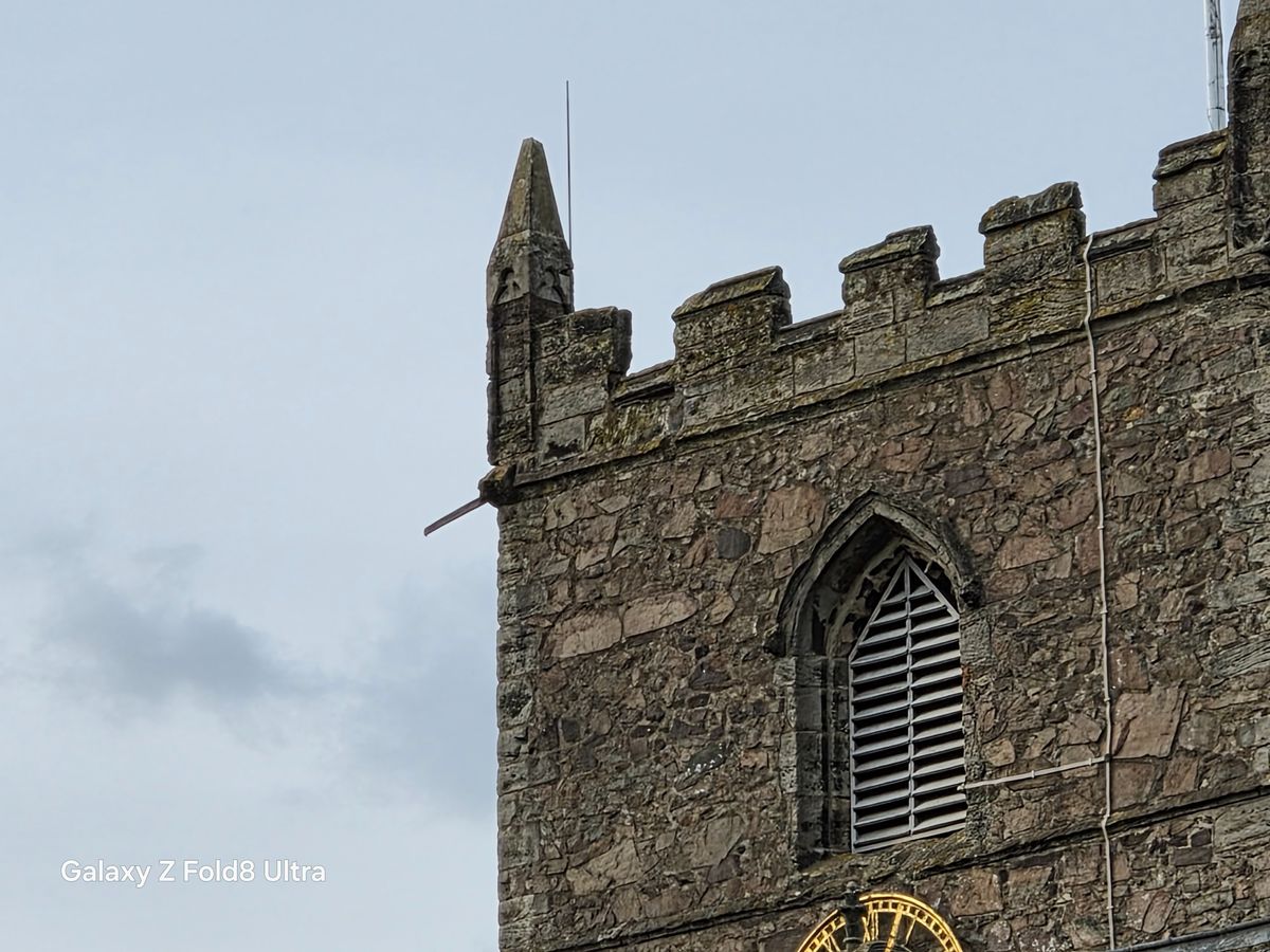 Close telephoto view of the upper section of a weathered stone church tower with a Gothic louvered window.