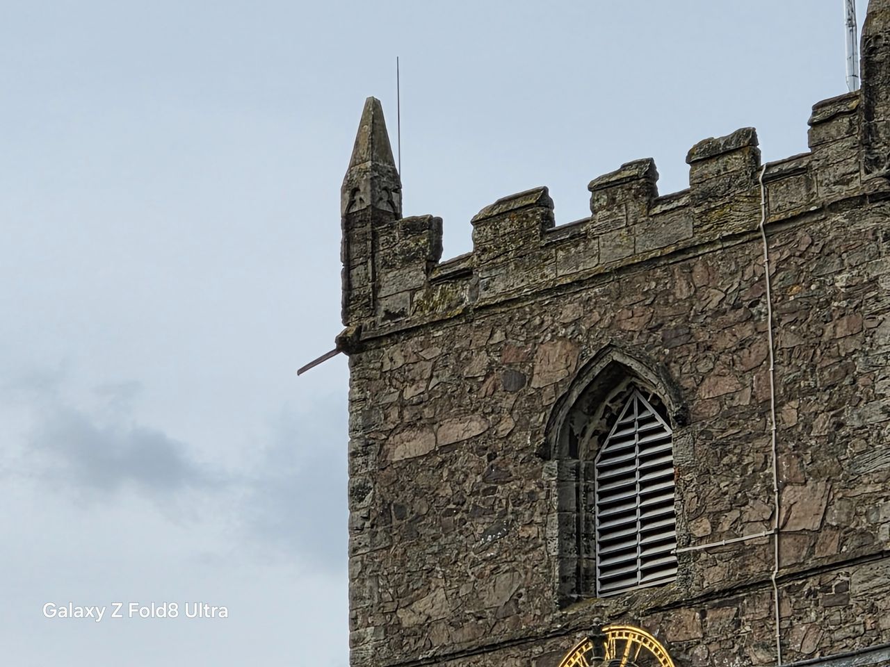 Close telephoto view of the upper section of a weathered stone church tower with a Gothic louvered window.