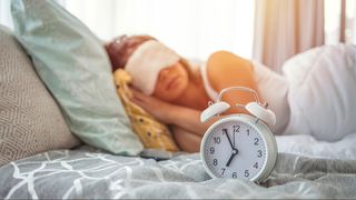 A woman sleeping on her side with eye mask on next to white alarm clock,