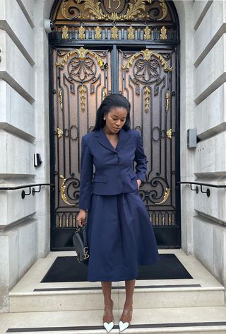 A woman standing in a doorway wearing a navy blue blazer with a matching skirt, white pumps, and a shoulder bag.