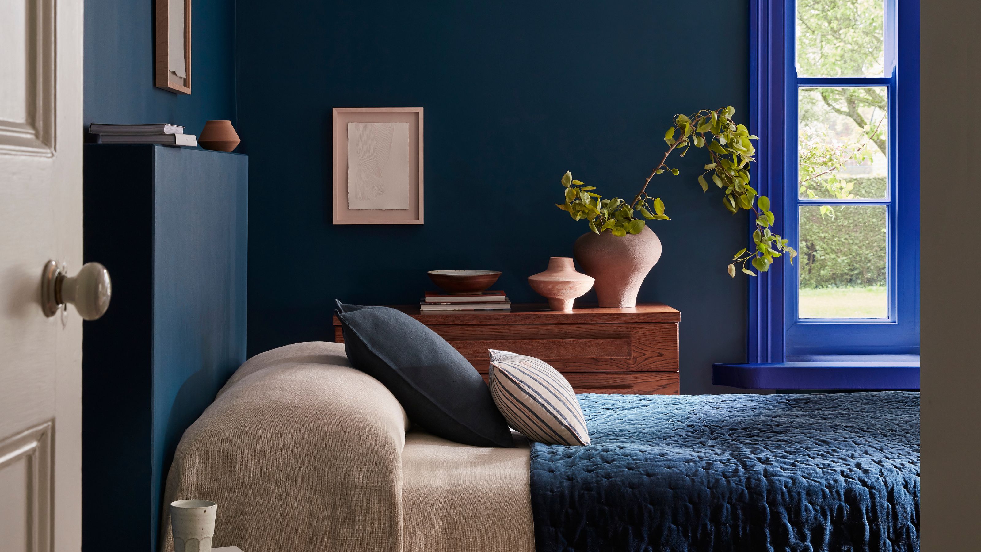 A dark blue bedroom with an electric blue window frame, a dark blue bedcover, and a navy blue nightstand. 