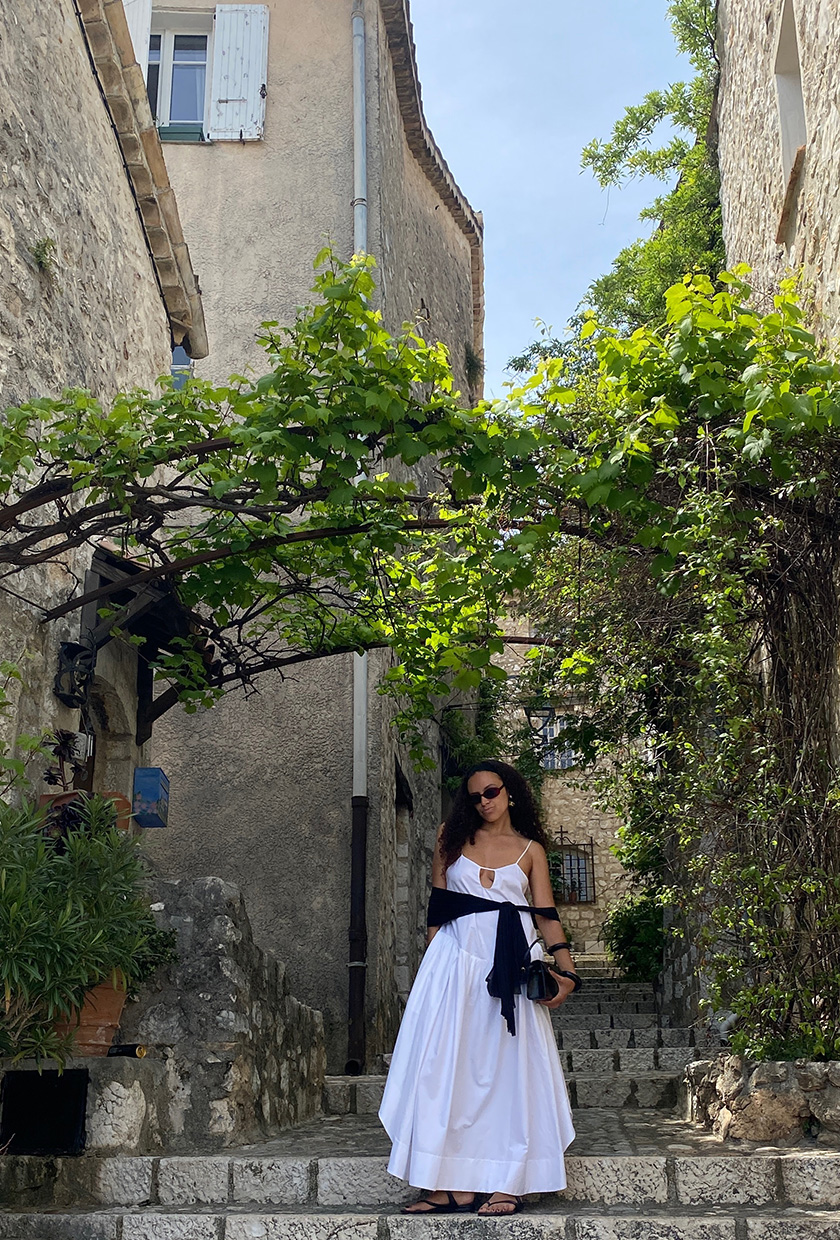 An editor standing on a street in Saint Paul de Vence wearing a white maxi dress, black scarf, black flip-flops, black bangles, and a black bag.