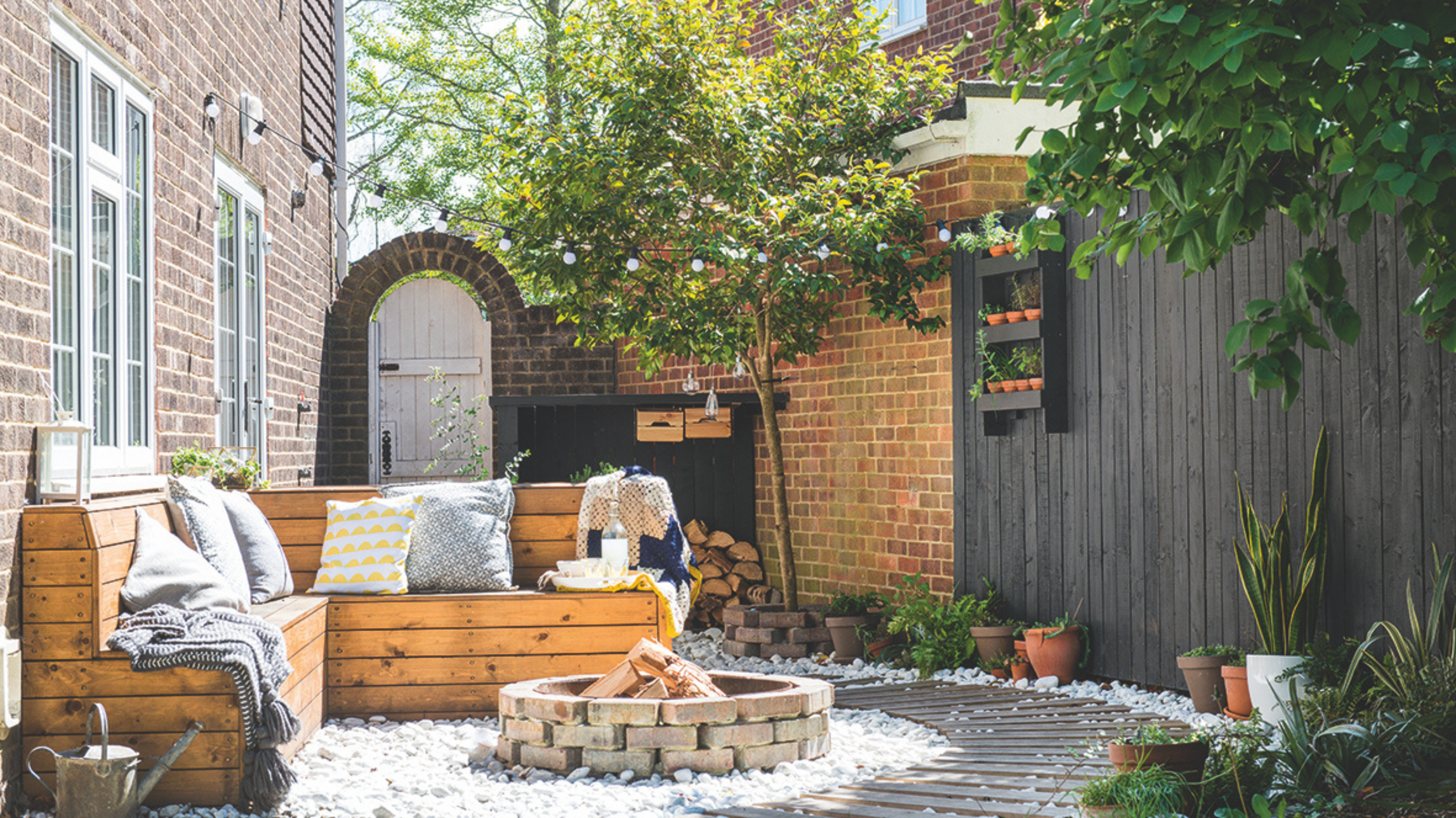Small garden with wooden outdoor seating, fire-pit and wooden boardwalk on white gravel, black wooden fence.