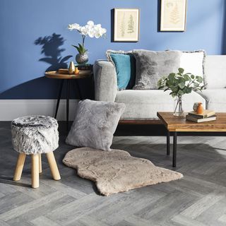 A blue living room with a grey sofa and wooden coffee table with a rug, cushions and stool from Aldi Cosy collection