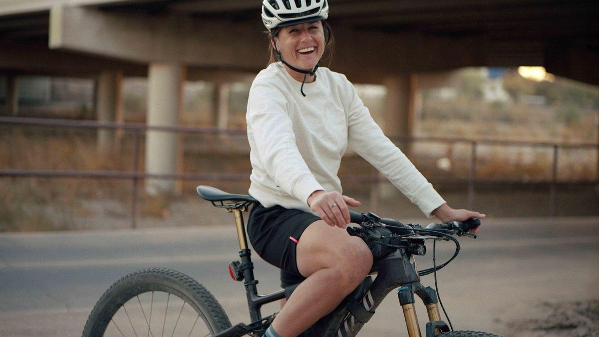 Lael Wilcox is sitting on her bike, wearing a white jumper, white helmet and black shorts, and is smiling at the camera