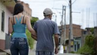 A woman wearing a blue tank top and jeans with long dark hair holds the arm of an older tan man wearing a gray shirt and a baseball cap as they walk down the street, their backs to the camera