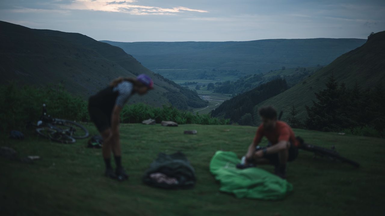 Two people layout bivy bags on grass overlooking a valley