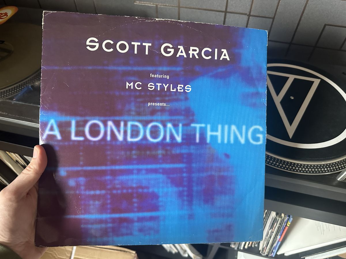 A hand holding a copy of Scott Garcia's A London Thing on vinyl. The cover is mostly blue and a turntable is visible in the background.