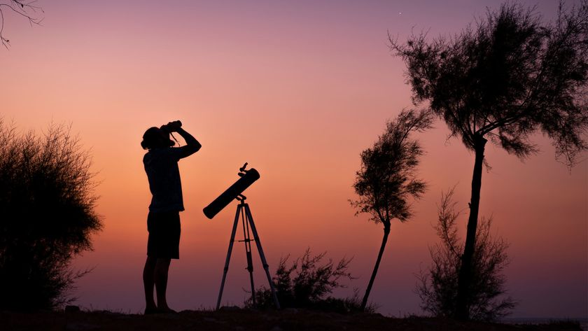 A man using binoculars and a telescope to view the sky. The sky is orange and purple, with the picture framed by trees and bushes.