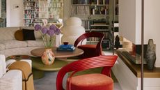 A modern, eclectic style living room with exposed beamed ceilings. white walls and large book cases on the two far walls. The seating area is comprised of one large curved sofa, two red armchairs and a multi level coffee table.