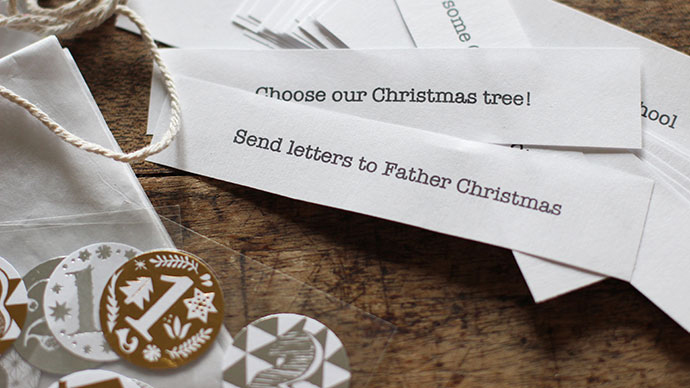 Wooden table with a pile of slips of white paper printed with advent activities next to a pile of festive numbered stickers