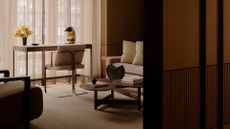 A minimalist study with a chair and desk by the window with a lamp and a vase of flowers. Behind the chair is a sofa, a coffee table with a vase and a book, and a plush sofa seater.