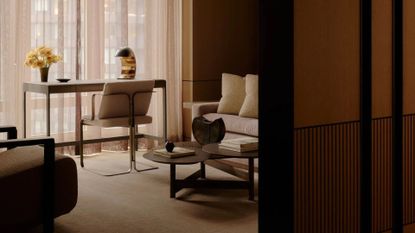 A minimalist study with a chair and desk by the window with a lamp and a vase of flowers. Behind the chair is a sofa, a coffee table with a vase and a book, and a plush sofa seater.
