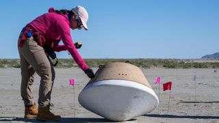 a woman in a baseball cap examines a mock sample-return capsule in a desert landscape.
