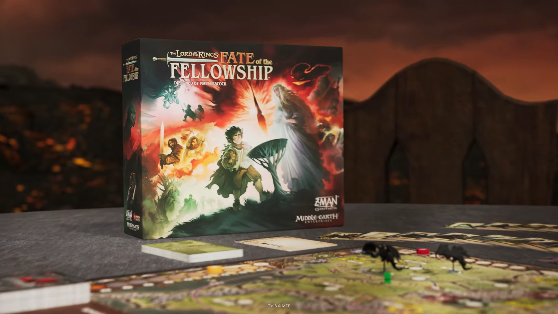 The box, board, cards, and tokens of Fate of the Fellowship set up on a slate-gray surface, against a volcanic background