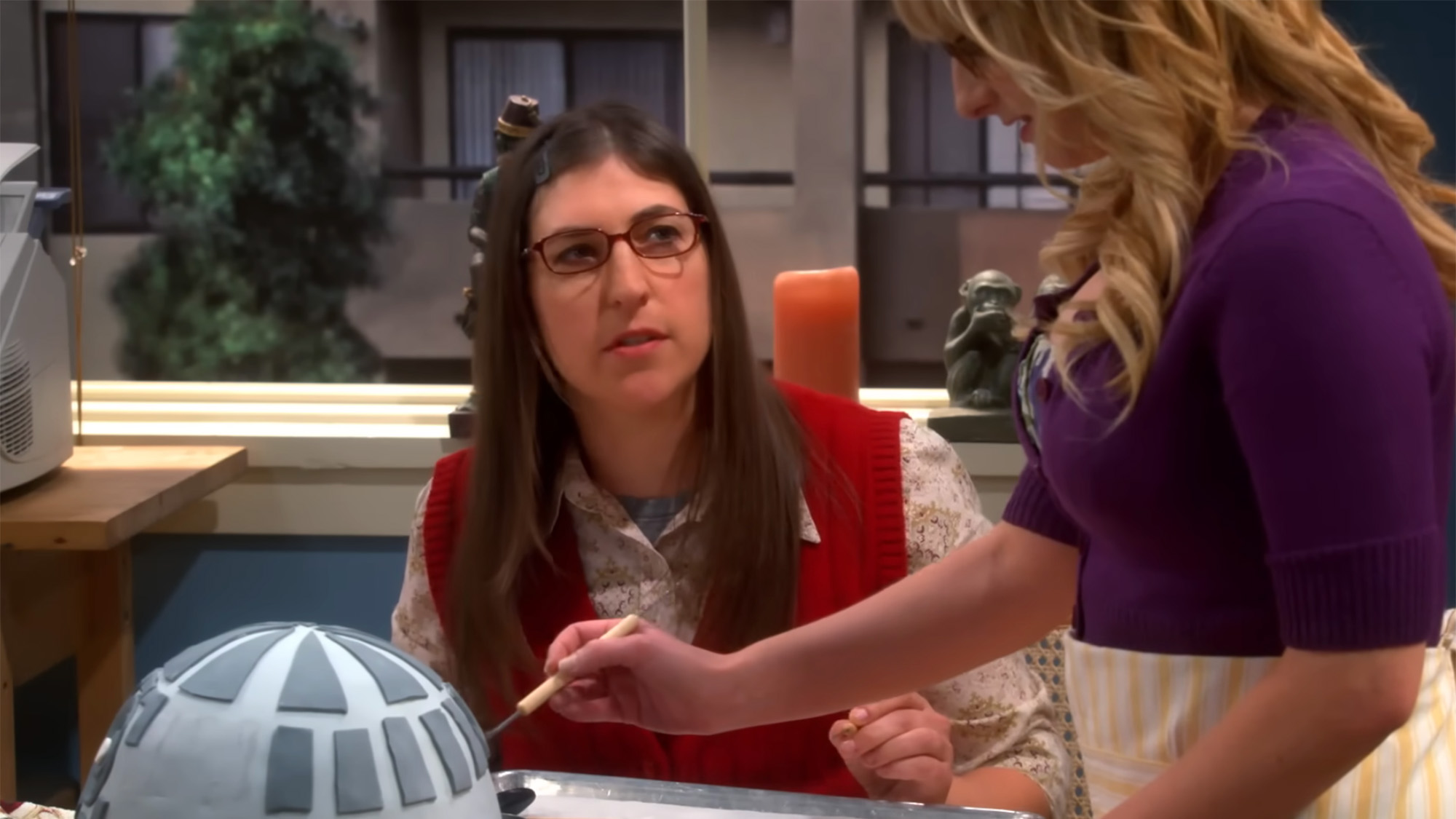 Mayim Bialik in a red sweater vest and glasses playing Amy Farrah Fowler on The Big Bang Theory. She is decorating a cake with Bernadette.