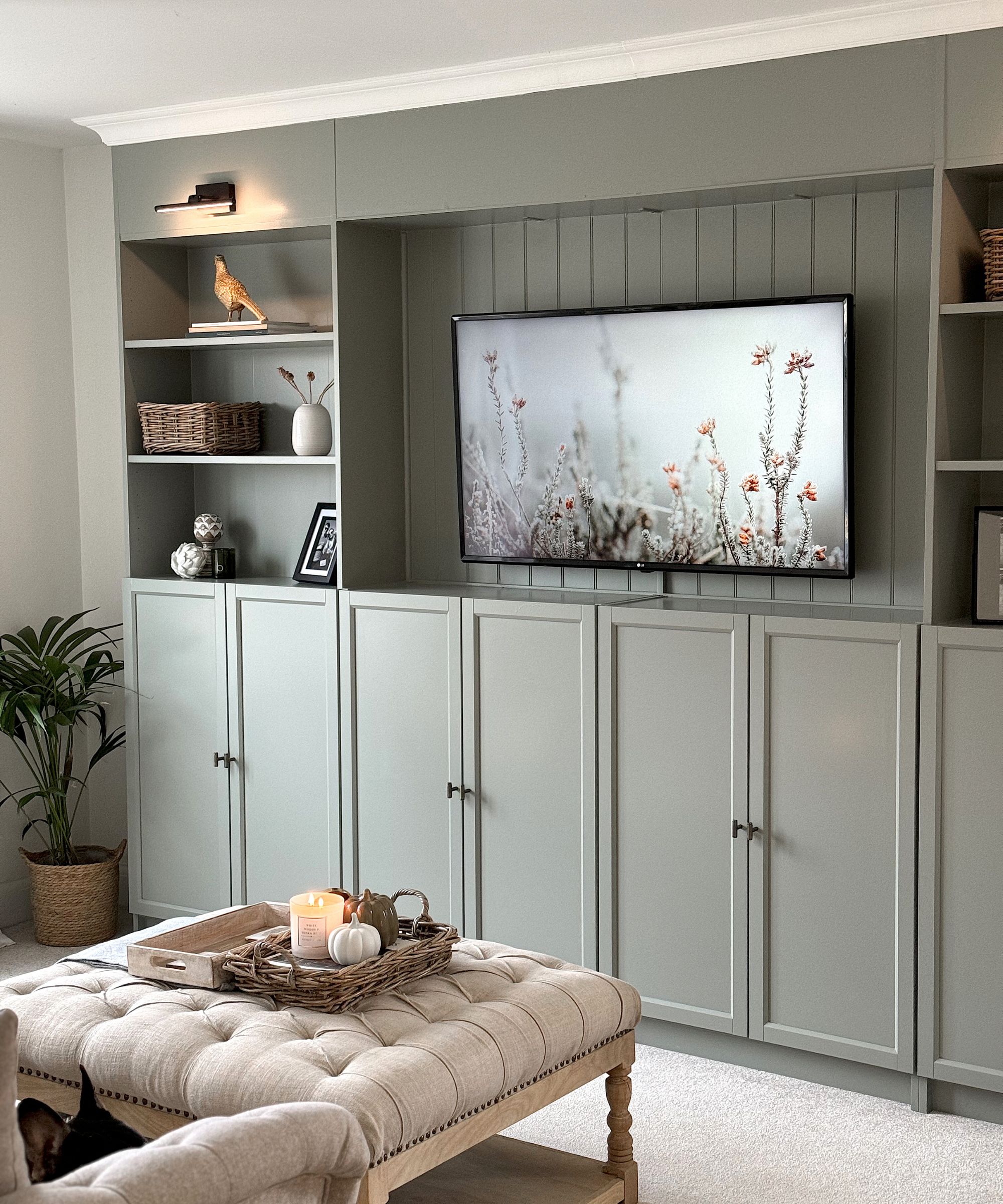 Living room with built-in grey storage, upholstered ottoman, shelves and rattan plant pot with plant