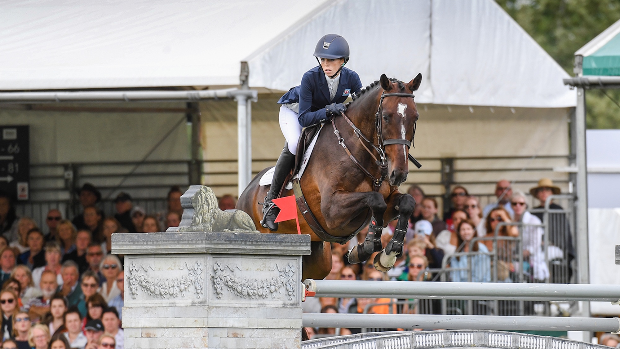 Image shows Alice Casburn (GBR) riding bay horse Topspin and jumping a grey showjump, away from crowds in a grandstand, at Burghley Horse Trials in 2022.