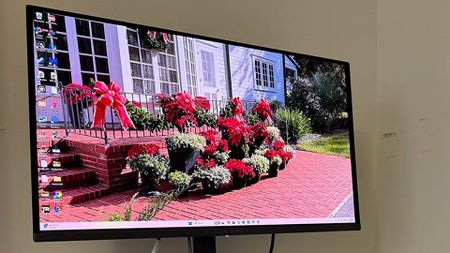 A computer monitor displays a bright desktop wallpaper of a white house, red brick steps and path, and vivid flowers in a sunny garden.