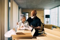 An older couple unwraps moving boxes and smile with the man's arm around the woman in a new home.