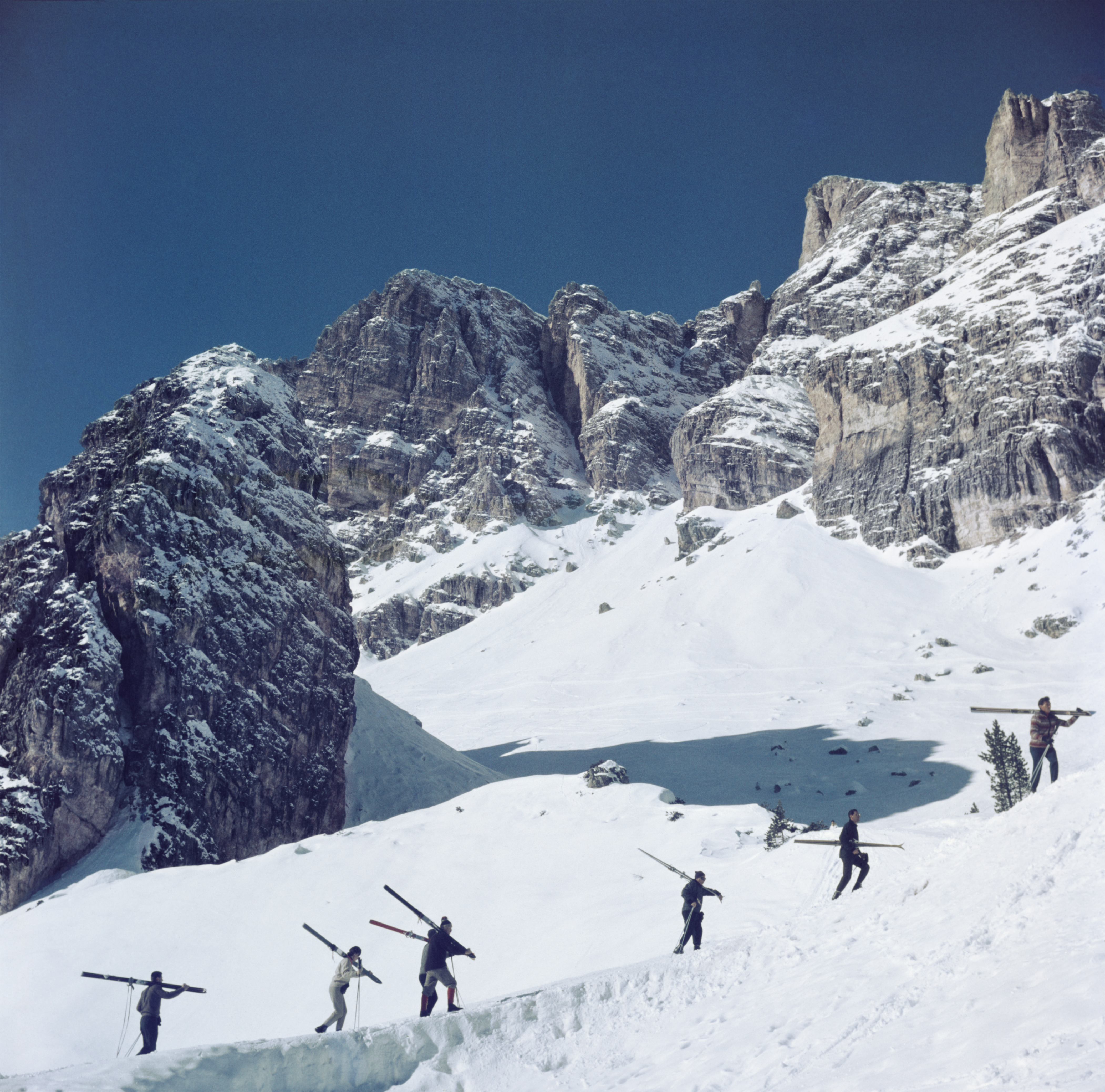 Skiers crossing a snow ridge beneath towering, jagged Dolomite peaks, carrying skis on their shoulders against a deep blue winter sky.