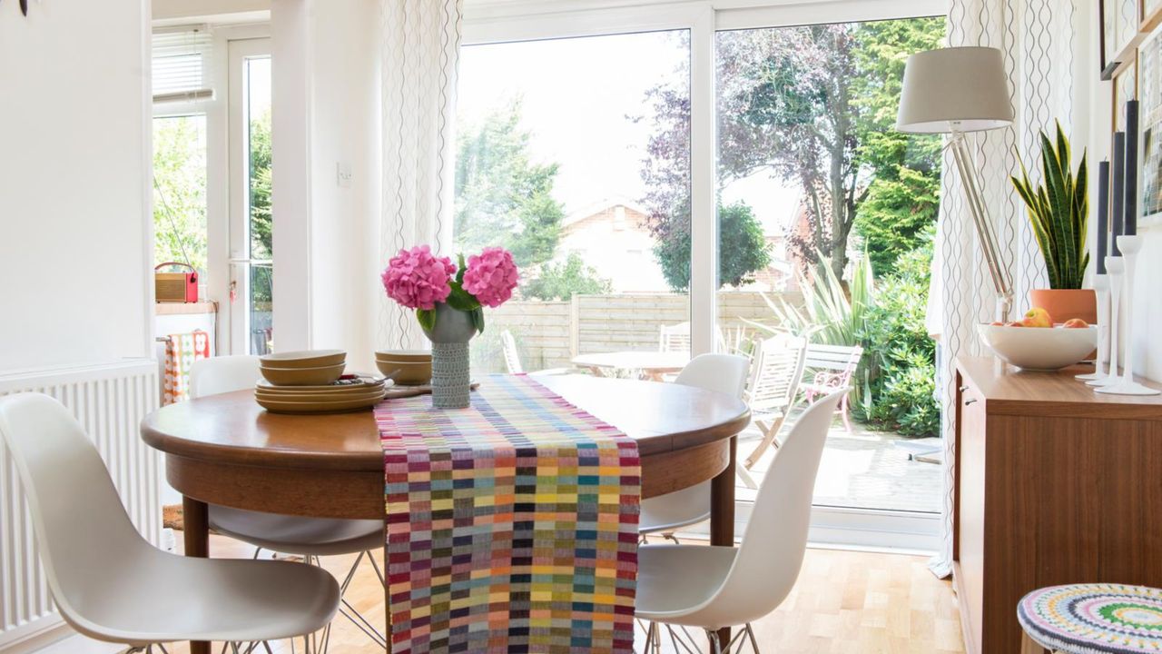 We love small dining room table ideas. Here&#039;s a dining room with a round dining table with a colorful table runner and pink flowers on it, a white chair, a wall with a glass window door looking onto a garden, a white lamp, a wooden storage unit with a plant and fruit bowl on, and a colorful stool