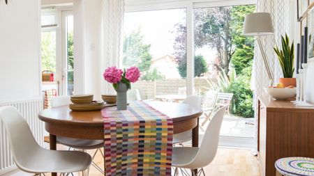 We love small dining room table ideas. Here's a dining room with a round dining table with a colorful table runner and pink flowers on it, a white chair, a wall with a glass window door looking onto a garden, a white lamp, a wooden storage unit with a plant and fruit bowl on, and a colorful stool