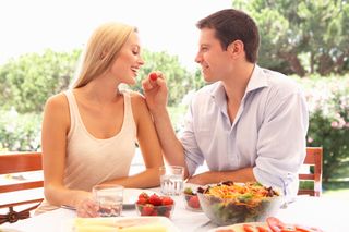 A couple sits at a table, the man holding up food for the woman to eat.